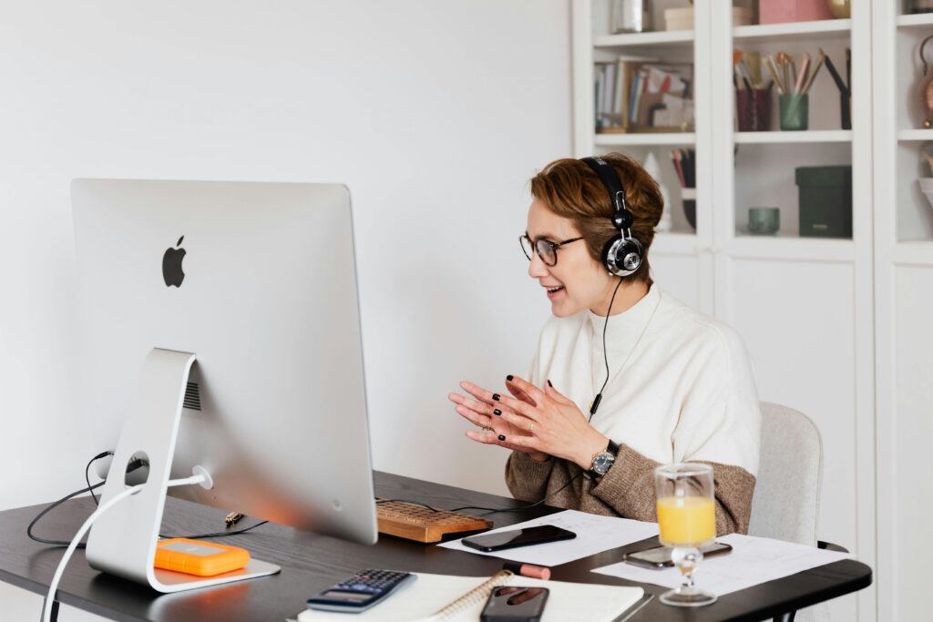 Cheerful female in casual wear and wired headphones having conversation via video call on computer while working in light modern workplace